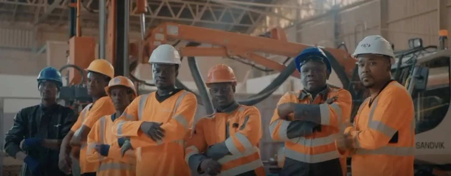 Huit membres d'une équipe de forage en tenue haute visibilité orange et casques de couleurs différentes, alignés bras croisés devant une foreuse Sandvik, sous un hangar d'atelier.