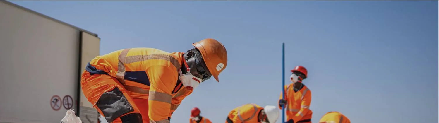 Un artificier en tenue haute visibilité orange, casque et masque de protection, se penche sur une charge au sol ; d'autres membres de l'équipe travaillent en arrière-plan sous un ciel dégagé.