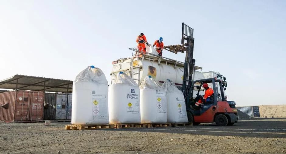 Aire de stockage sous hangar : des grands sacs blancs de produit alignés sur des palettes, un chariot élévateur rouge et un bâtiment technique.