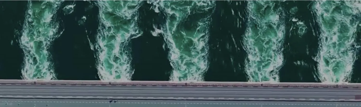 Vue aérienne d'un barrage : l'eau s'écoule par plusieurs vannes en formant de larges panaches d'écume blanche sur une eau vert sombre.